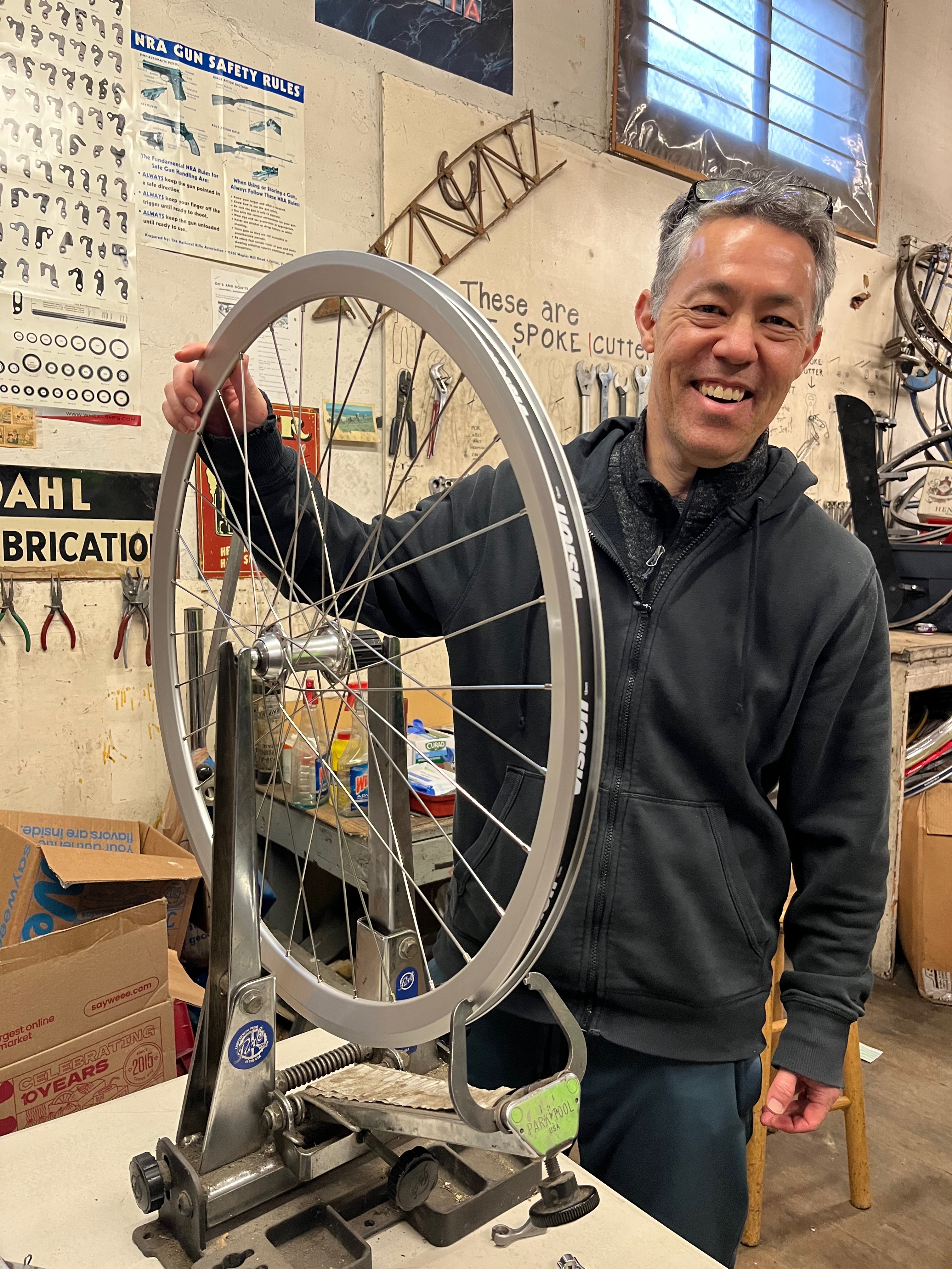 Jon Wren smiling beside a freshly built bicycle wheel on a truing stand in a bike shop